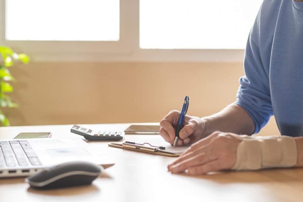 Person with a wrist brace writing on a clipboard at a desk with a laptop and calculator.